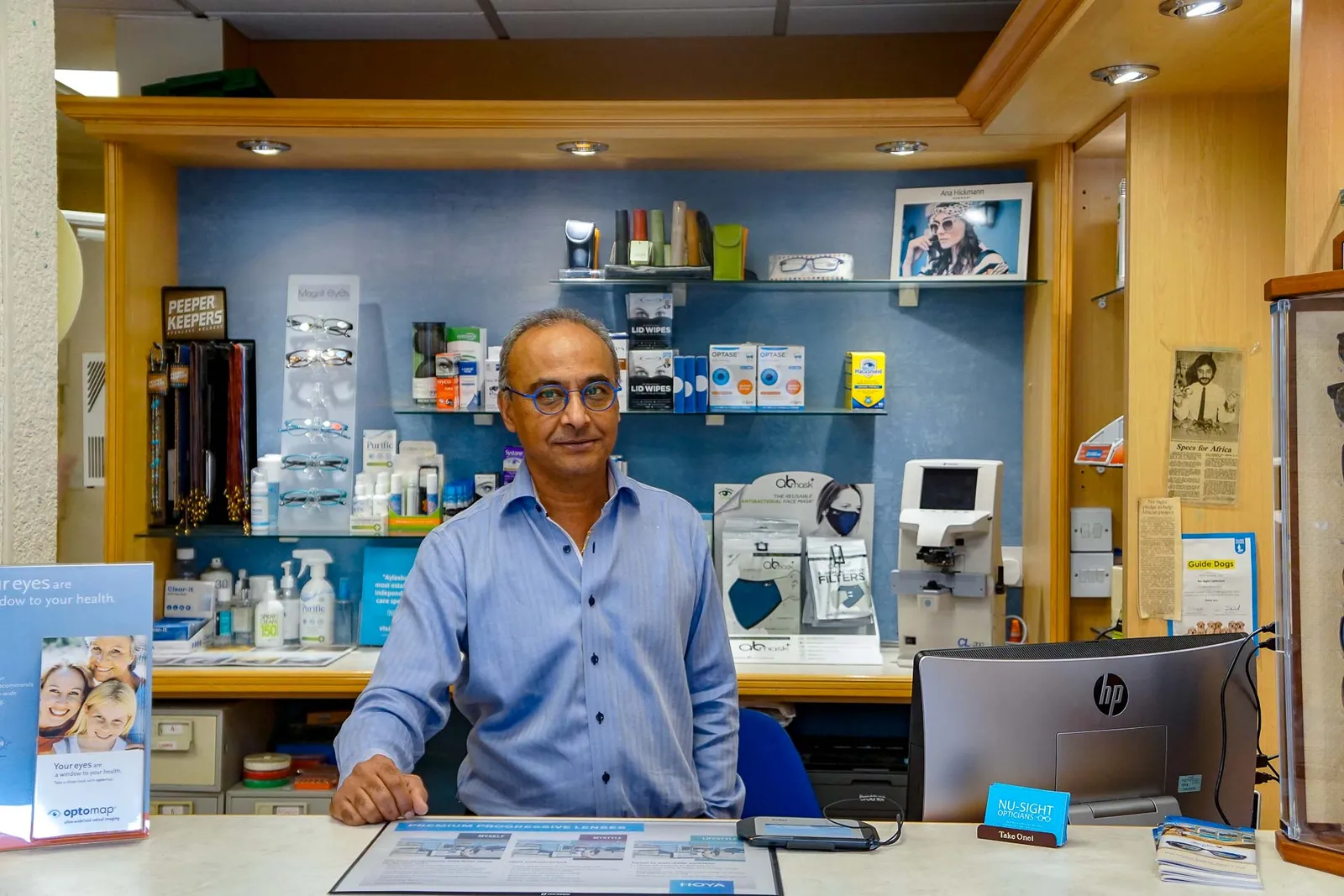 An optometrist at the dispensing desk inside Nu-Sight Opticians, with frames and lens displays behind